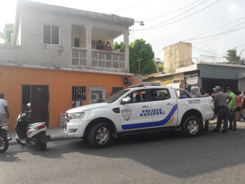 Vehículo de la Policía Nacional estacionado frente a una vivienda en La Romana durante un operativo, mientras curiosos observan la escena.